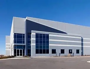 A large, modern industrial building with gray and white exterior walls, multiple large windows, and a paved area in front under a clear blue sky.